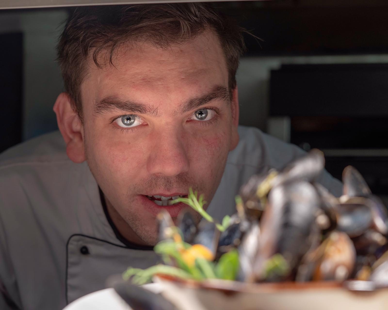 A chef holding a bowl of mussels towards the camera in the kitchen.
