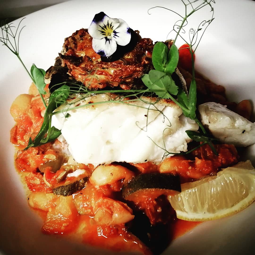A white fish fillet on a tomato sauce with mussels, vegetables and a lemon wedge.